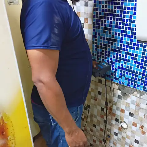 Técnico com geofone encostado na parede de pastilhas de um banheiro, ouvindo a água na tubulação embutida